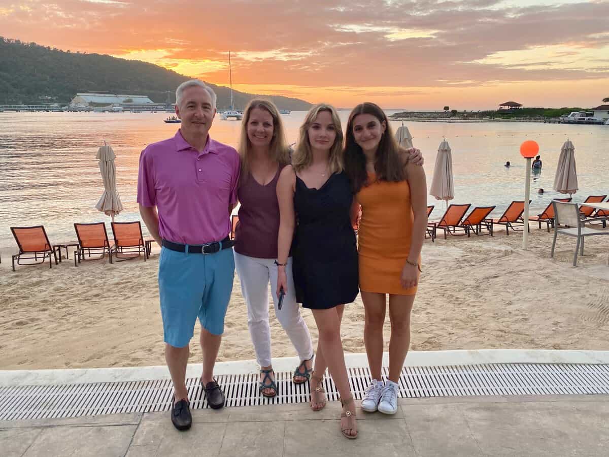 A family of four poses together on the beach at Moon Palace Jamaica, standing in front of lounge chairs and umbrellas with the golden sunset reflecting on the calm water behind them. The group smiles warmly with the ocean, sailboats, and hills forming a scenic tropical backdrop.