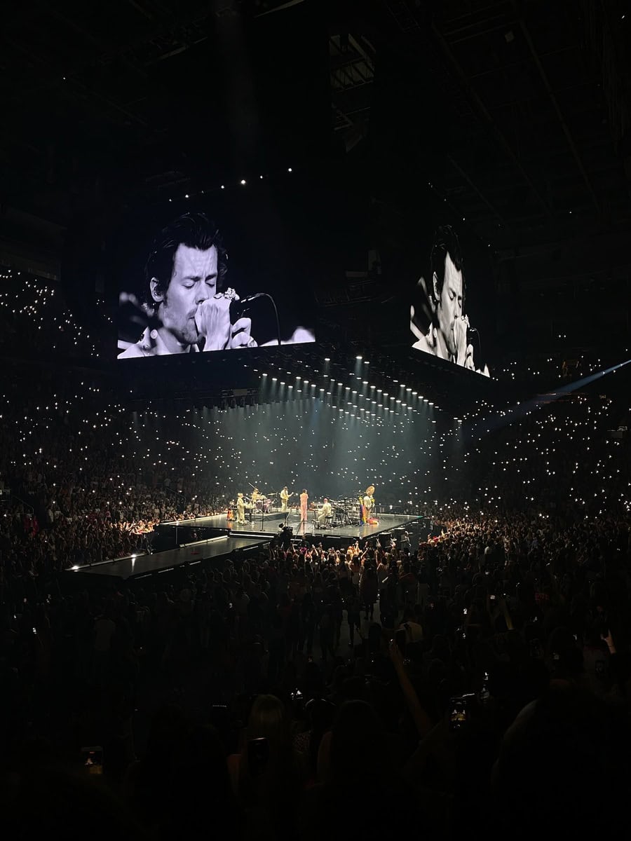 Harry Styles performs in a packed arena in Toronto, standing on a central stage surrounded by fans, with thousands of phone lights glowing in the dark. A giant screen overhead shows a black-and-white close-up of him singing emotionally into the microphone.