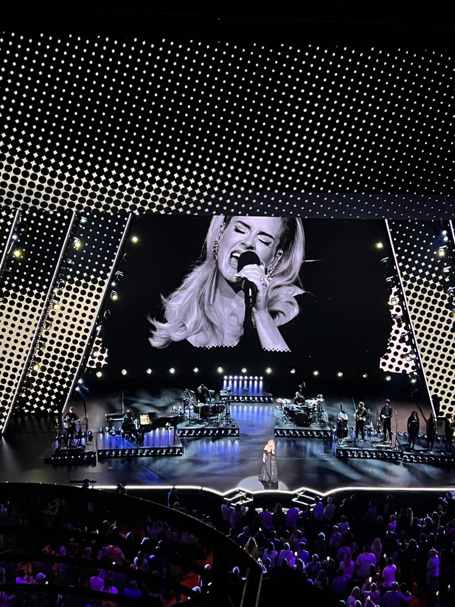 Adele performs onstage in a long gown at The Colosseum in Las Vegas, with a black-and-white close-up of her singing projected on a massive screen behind her. The stage is framed by a dotted arc of lights and a full band setup, with a captivated audience in front.