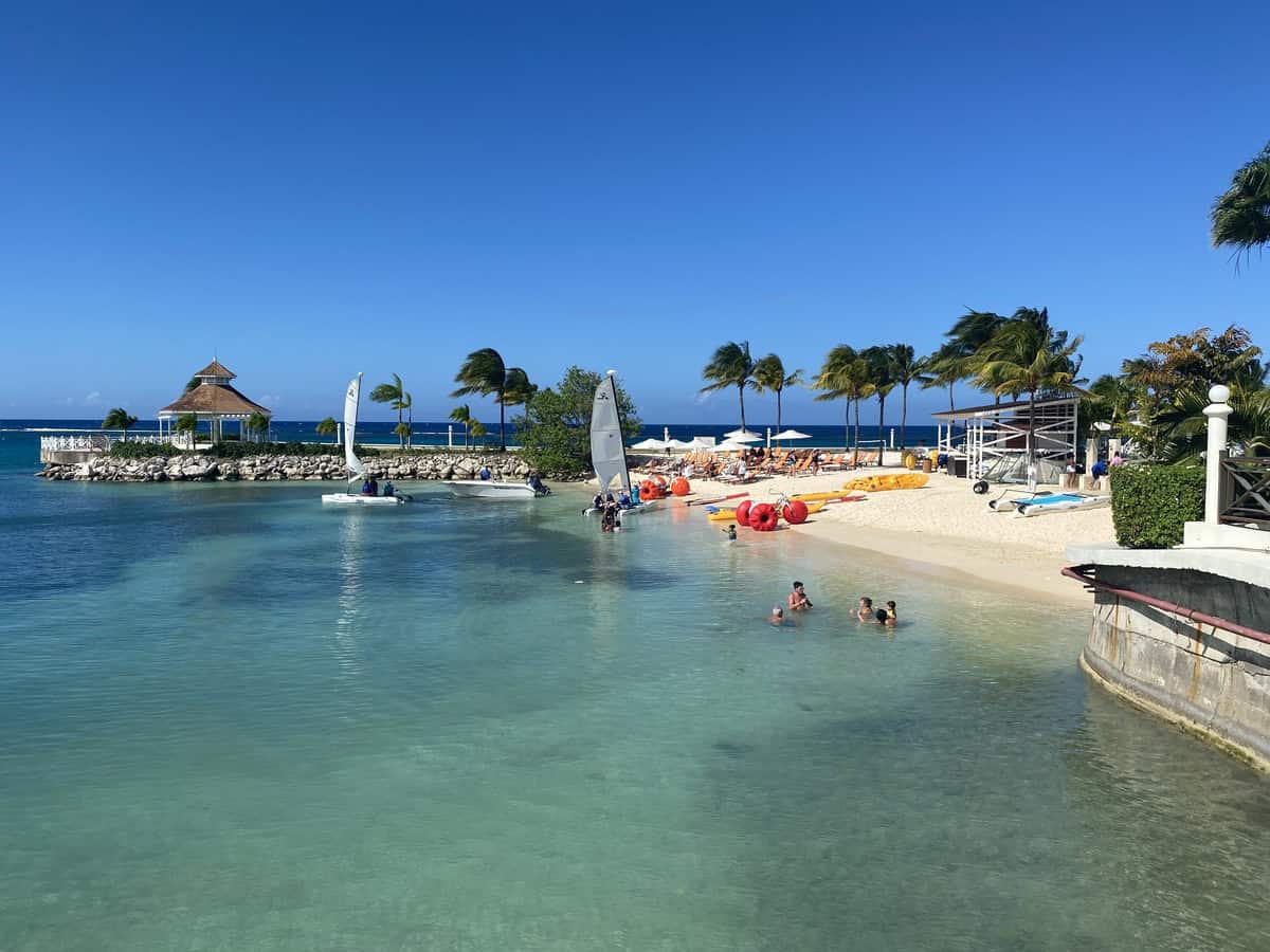 Guests enjoy a day of water sports at Moon Palace Jamaica, with sailboats, kayaks, and paddleboards lined up on the sandy beach. Clear turquoise water surrounds a small gazebo-topped island and rows of loungers shaded by palm trees.