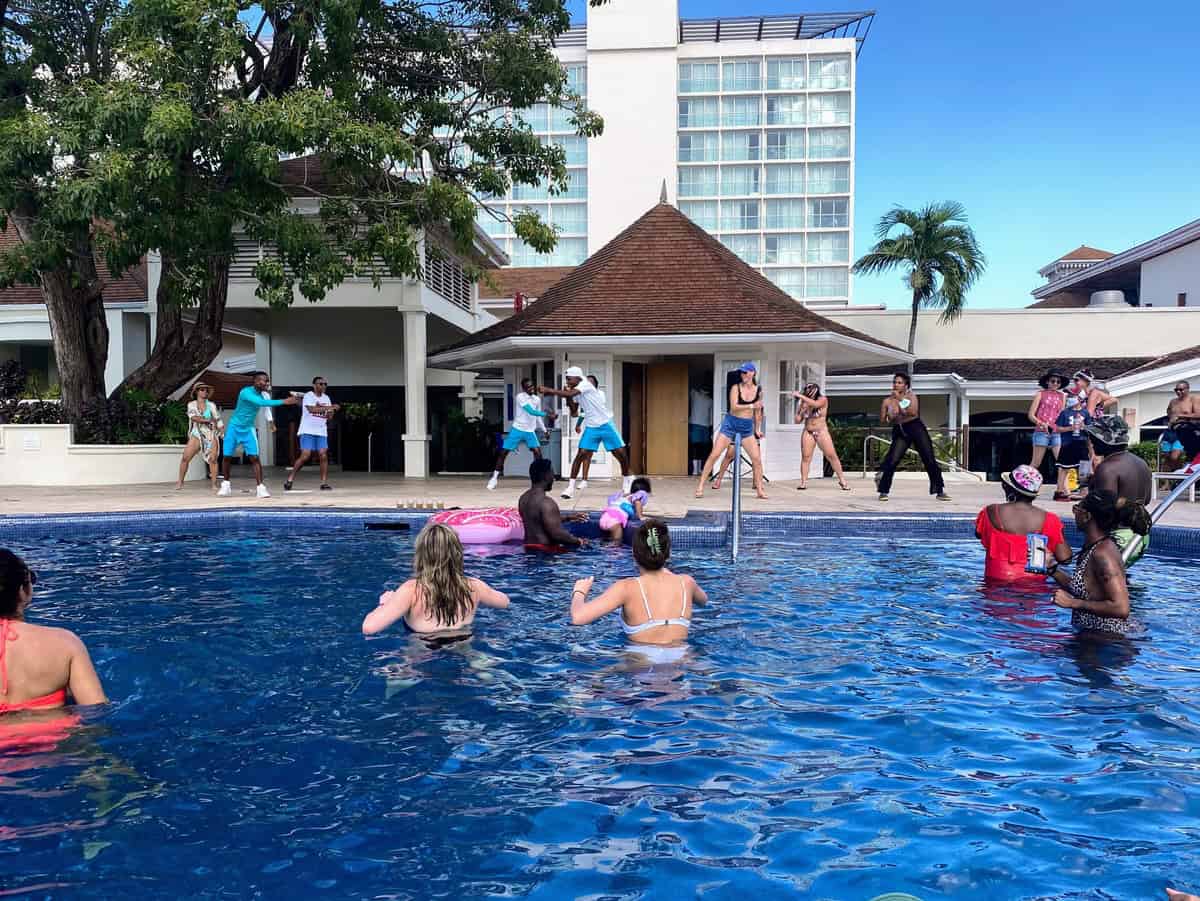 A lively poolside scene at Moon Palace Jamaica where guests in swimwear participate in a dance or exercise session led by energetic staff in turquoise outfits. Spectators and participants enjoy the activity under the tropical sun, with the modern resort buildings in the background.