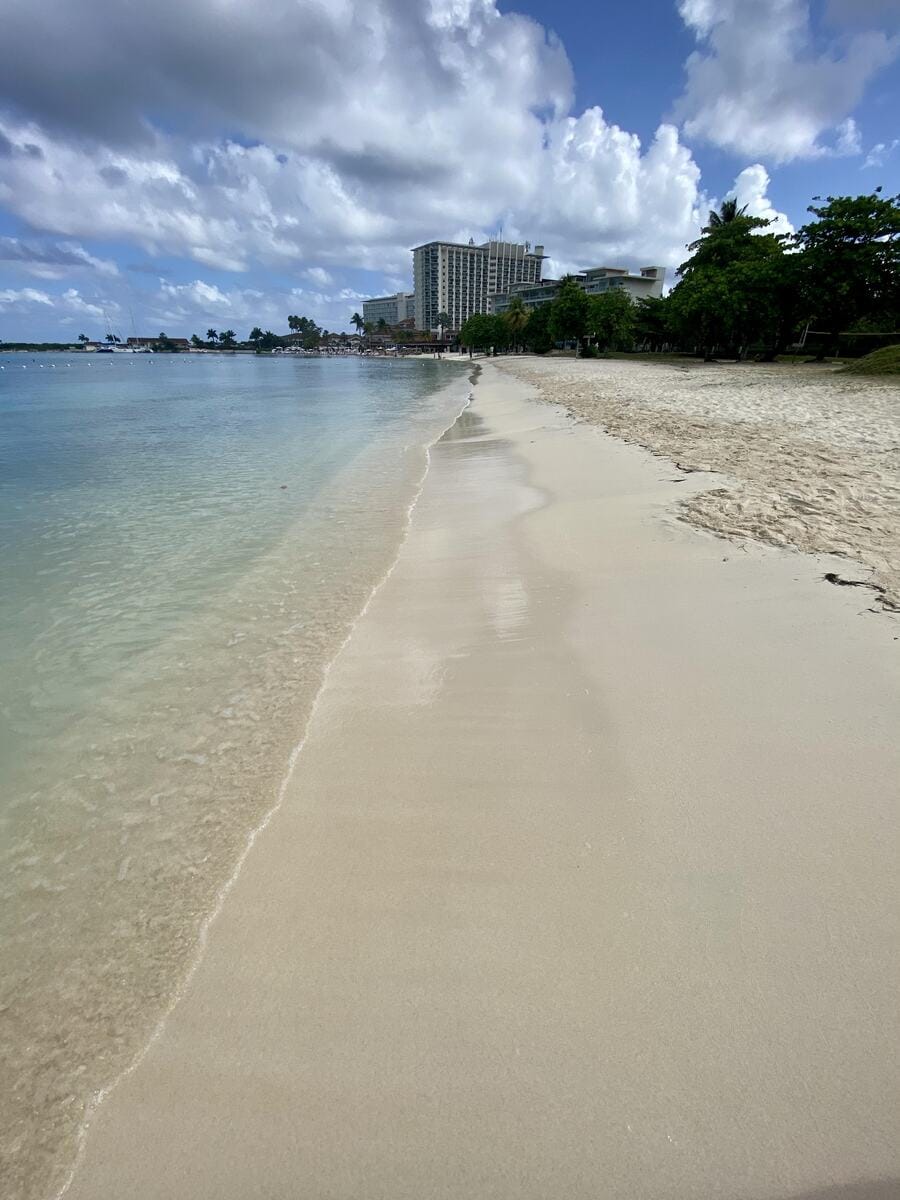 A peaceful stretch of soft sandy beach leads toward the Moon Palace Jamaica hotel, visible in the distance along the shoreline. The calm, clear water gently laps the shore under a sky filled with dramatic, fluffy clouds, with lush greenery lining the right side of the frame.