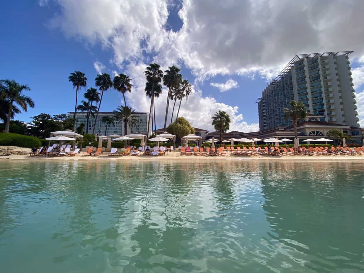 A peaceful view from the ocean looking toward Moon Palace Jamaica's beach, where orange lounge chairs and white umbrellas line the shore. Towering palm trees and the resort’s modern buildings complete the tropical scene.