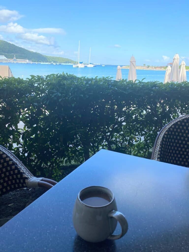 A ceramic mug of coffee rests on a sleek black table at Pier 8 restaurant, with a scenic view of sailboats anchored in the calm blue water. Green shrubs and umbrella-shaded lounge chairs frame the tranquil oceanfront backdrop.