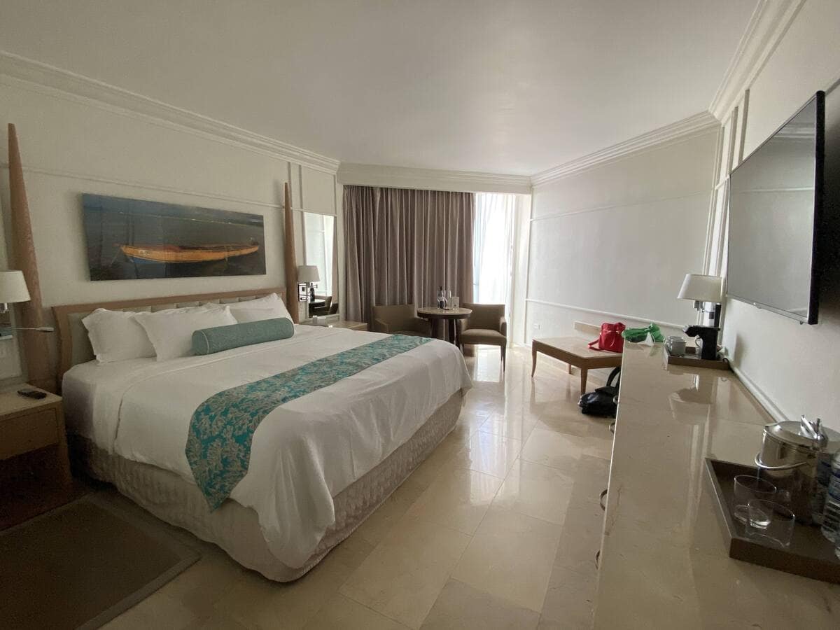 A cozy, well-appointed guest room at Moon Palace Jamaica featuring a large king bed with turquoise accents, a flat-screen TV, and a sitting area near a window with natural light filtering through. Modern furnishings and a clean design create a relaxing ambiance.