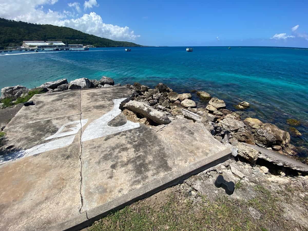 A cracked concrete slab featuring a faded white silhouette of James Bond points toward the turquoise ocean at Moon Palace Jamaica. Surrounded by rocky shoreline and lush green hills in the distance, this scenic and weathered spot marks a subtle tribute to the island's cinematic history.