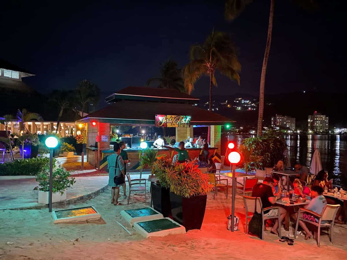 Guests dine and mingle at the beachside Jerk Hut under colorful lights at night. The open-air eatery glows with red and green lights while the dark water reflects city lights in the background, creating a festive Caribbean ambiance.