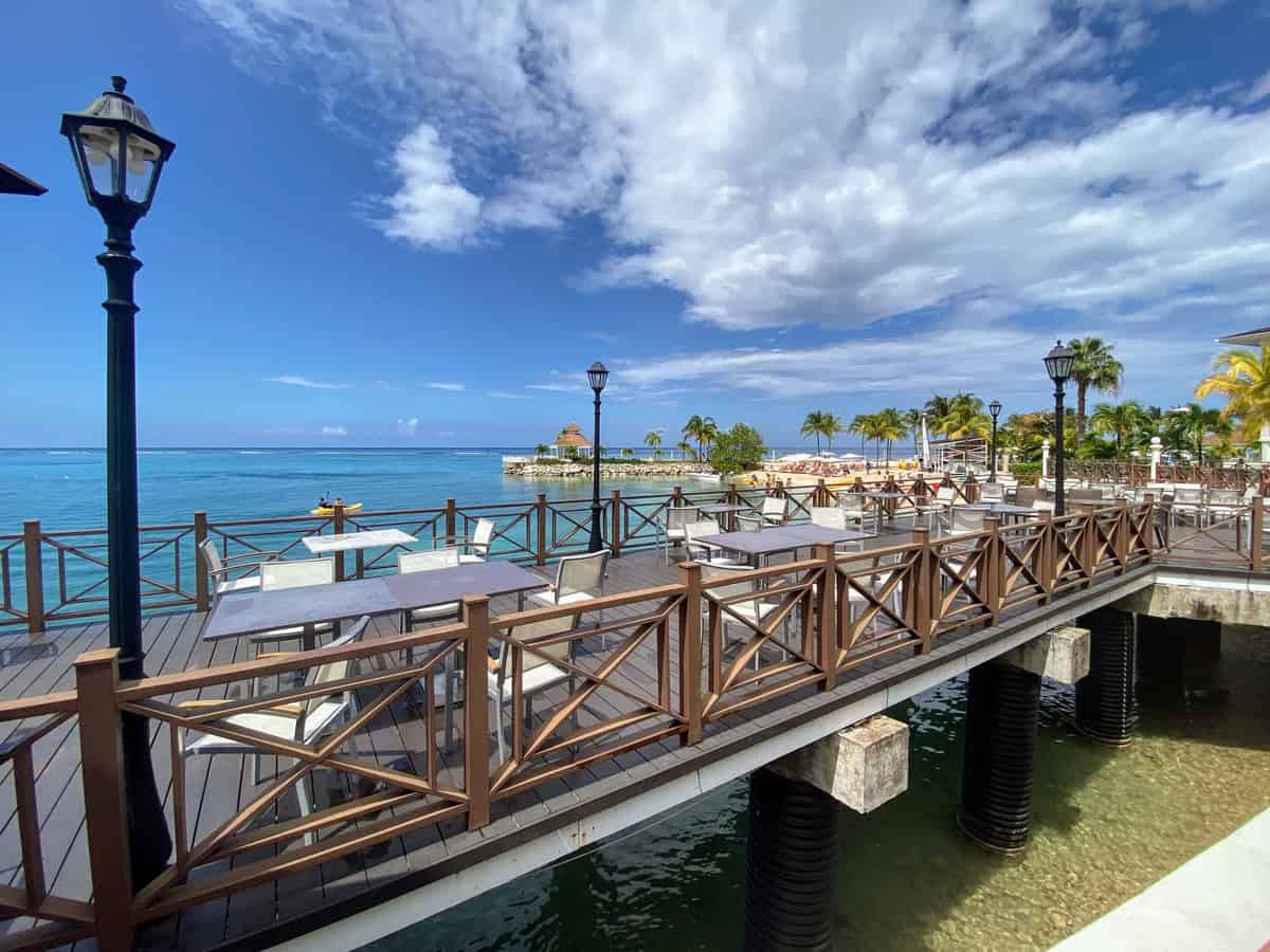 Outdoor dining area at Moon Palace Jamaica set on a wooden deck over the water, featuring white tables and chairs under classic lampposts. The view includes calm blue sea, distant palm trees, and a small island gazebo, all under a partly cloudy sky.