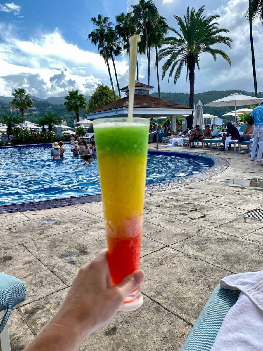 A hand holds a tall, tri-color frozen drink in red, yellow, and green, echoing the Jamaican flag, in front of a bustling pool at Moon Palace Jamaica. Guests relax and swim under bright blue skies with lush palm trees and mountains in the background, capturing a perfect tropical vacation vibe.