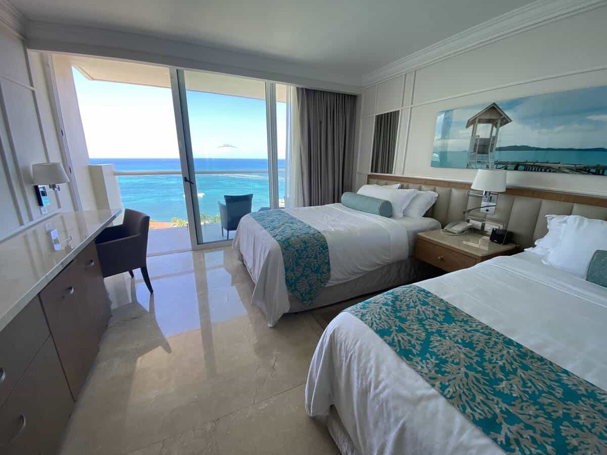 A bright, elegantly decorated hotel room in the Presidential Suite at Moon Palace Jamaica features two queen beds with turquoise coral-patterned runners. Floor-to-ceiling windows open to a balcony with a stunning view of the deep blue Caribbean Sea.