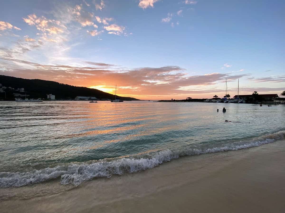 A tranquil sunset casts warm orange and pink hues across the sky and reflects on the gentle waves at Moon Palace Jamaica. Silhouettes of swimmers and anchored boats dot the peaceful water as the sun sets behind the hills in the distance.