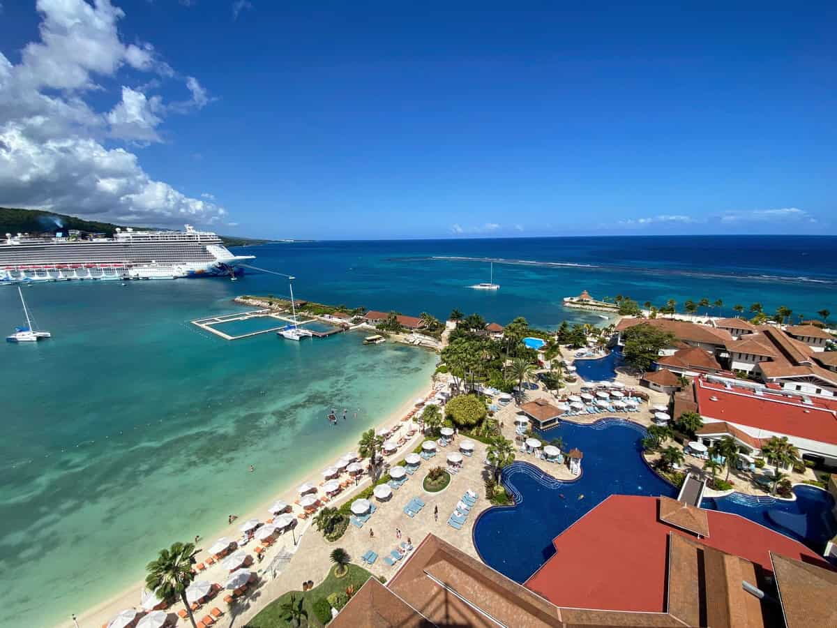A wide-angle aerial view of Moon Palace Jamaica captures the resort’s beachfront lined with umbrellas and lounge chairs, several curvy pools, and a cruise ship docked nearby. The vivid turquoise water stretches to the horizon under a clear blue sky.