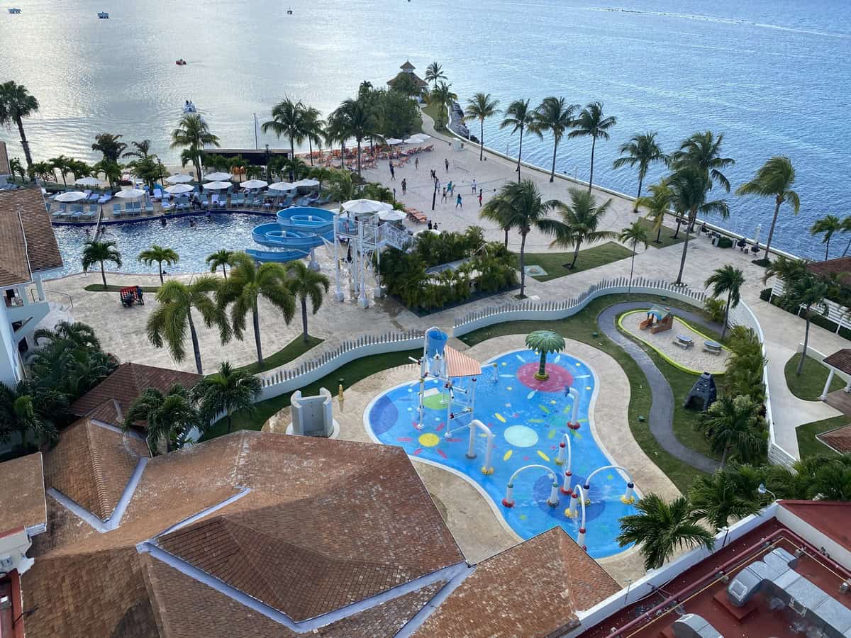 An aerial shot overlooking the pool complex at Moon Palace Jamaica, including a water play area for kids with colorful splash features, a waterslide tower, and adults lounging poolside near the oceanfront.