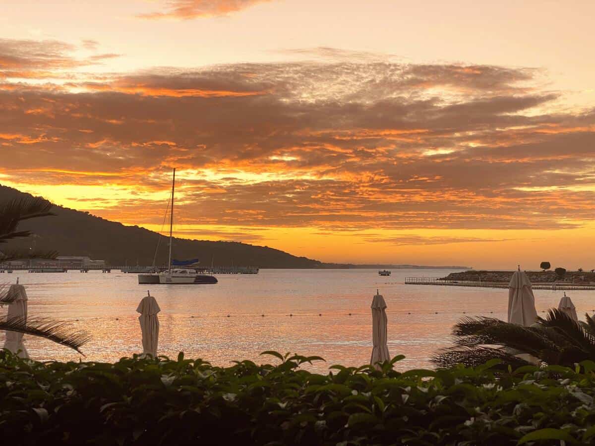 A breathtaking sunset bathes the sky in rich shades of orange and gold over the calm waters at Moon Palace Jamaica. A sailboat floats peacefully near the dock while closed umbrellas and lush greenery frame the foreground, creating a serene, postcard-worthy moment.