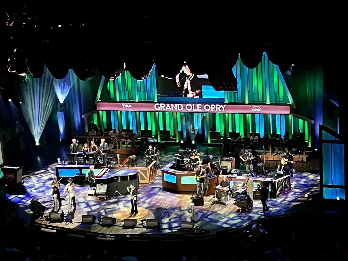 A full band and multiple performers play live onstage at the Grand Ole Opry in Nashville, Tennessee, with the signature wooden backdrop and “Grand Ole Opry” banner lit in green and blue. The classic country music venue features singers, guitarists, and a visible studio audience.