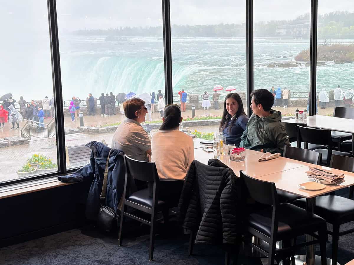 A group of four people sits at a window-side table inside Table Rock Restaurant, enjoying a meal with a stunning close-up view of Niagara Falls Canada and crowds of visitors watching the falls outside on a rainy day.