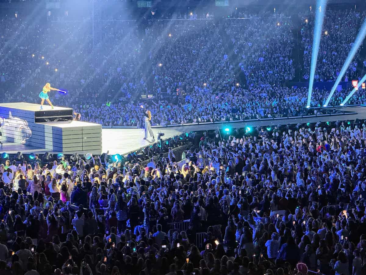 Taylor Swift performs on an elevated platform in a sparkling blue outfit during The Eras Tour in Toronto, with stage lights beaming and a massive, energetic crowd filling the Rogers Centre. Fans are on their feet with phones raised, capturing the moment under vibrant blue and purple lighting.