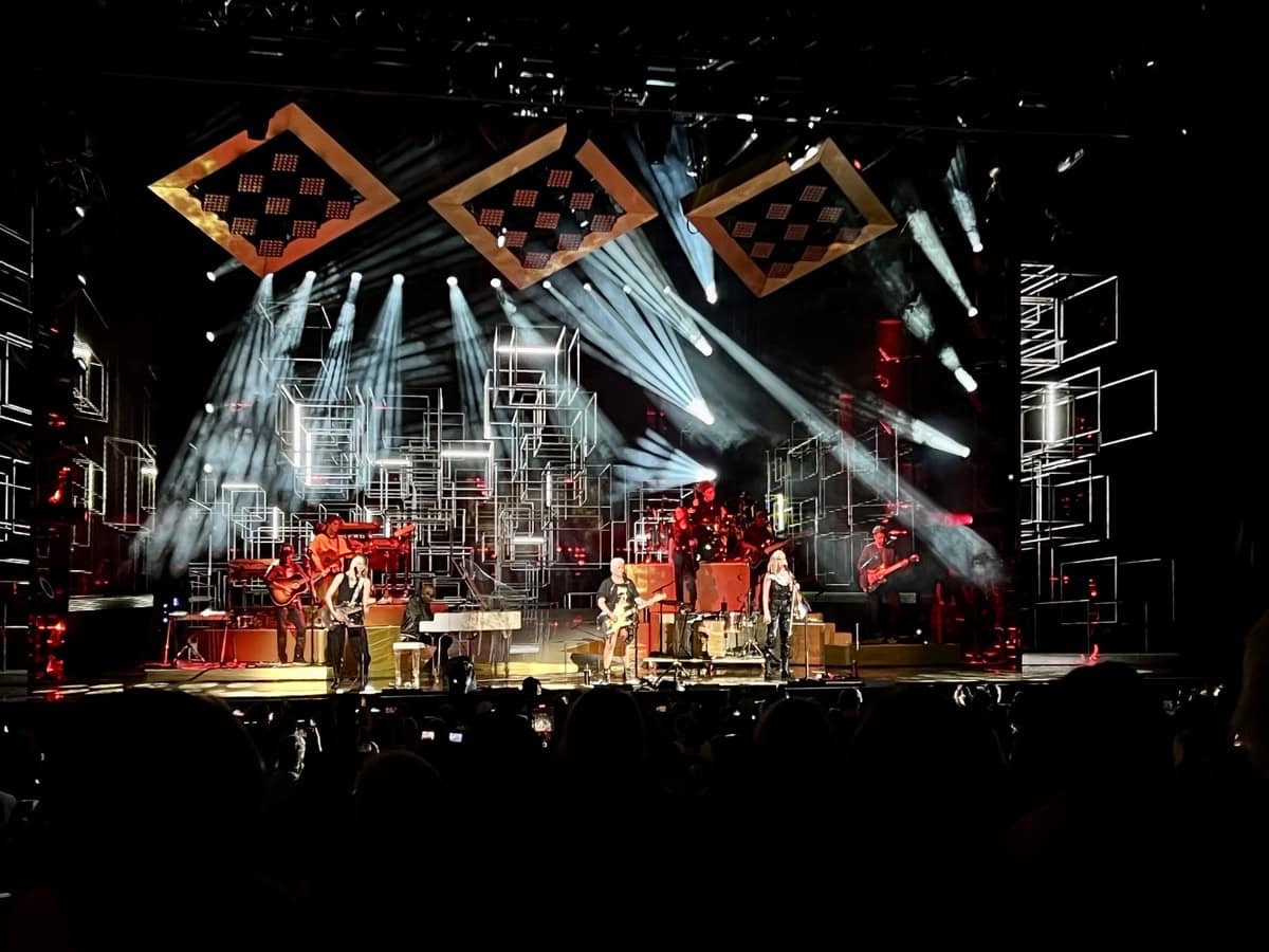 The Chicks perform onstage at Budweiser Stage in Toronto, backed by a full band and illuminated by white and red spotlights. The stage is framed by glowing geometric structures and suspended diamond-shaped lights, with a large audience visible in the foreground.