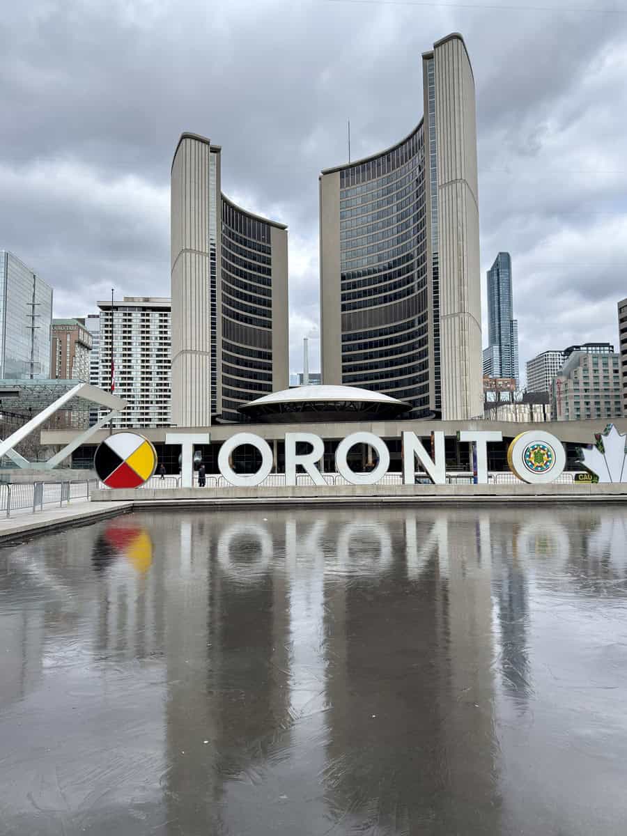 The iconic Toronto sign in Nathan Phillips Square is reflected on the surface of an empty skating rink, with the curved towers of City Hall standing tall under a moody, overcast sky.