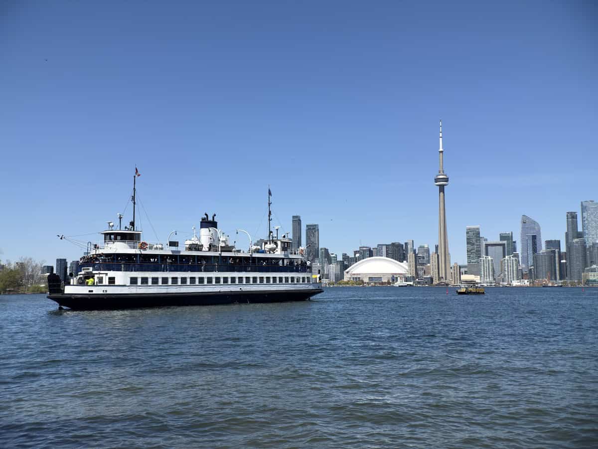 A Toronto ferry sails across the water with the city skyline in the background, featuring the CN Tower and Rogers Centre under a clear blue sky, as seen from Centre Island.