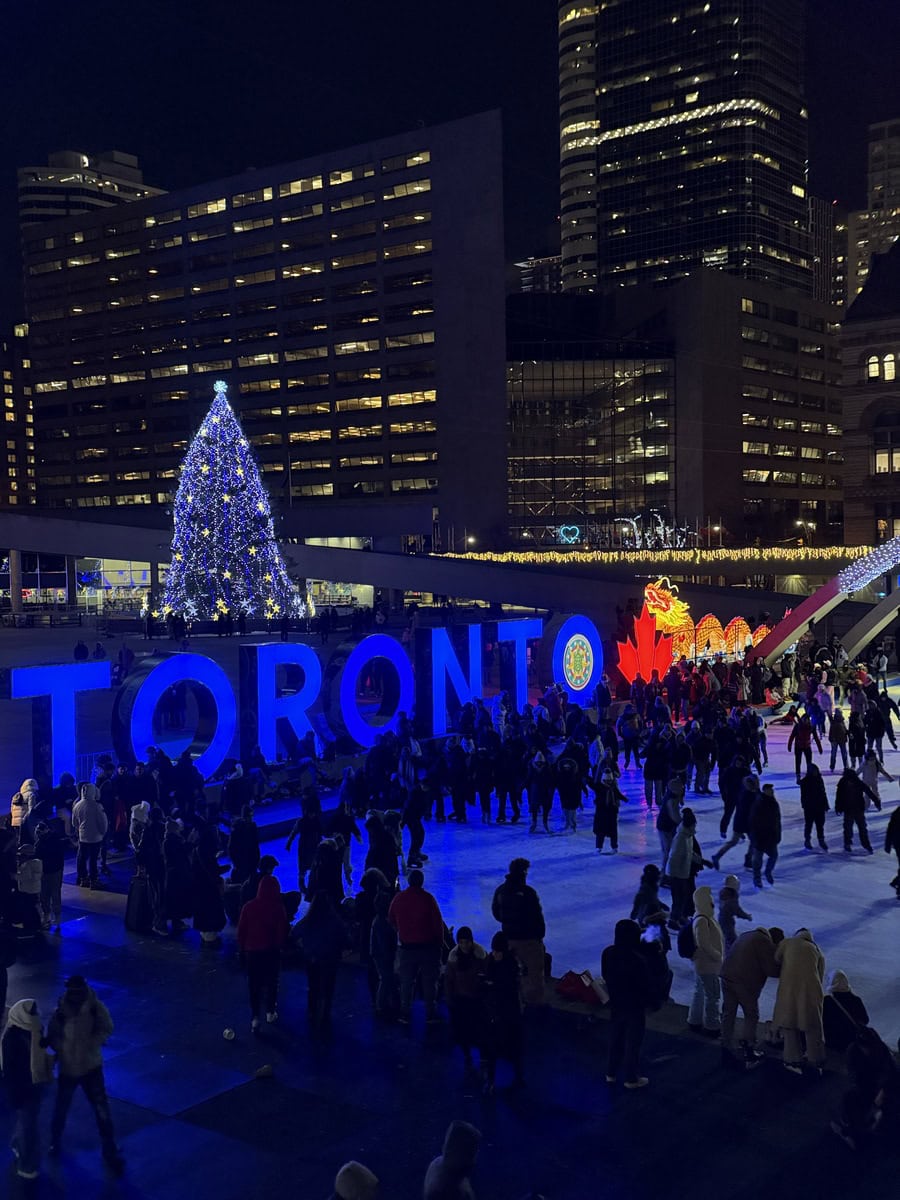 A crowd skates under glowing holiday lights at night in front of the illuminated Toronto sign and Christmas tree in Nathan Phillips Square, surrounded by high-rise buildings.
