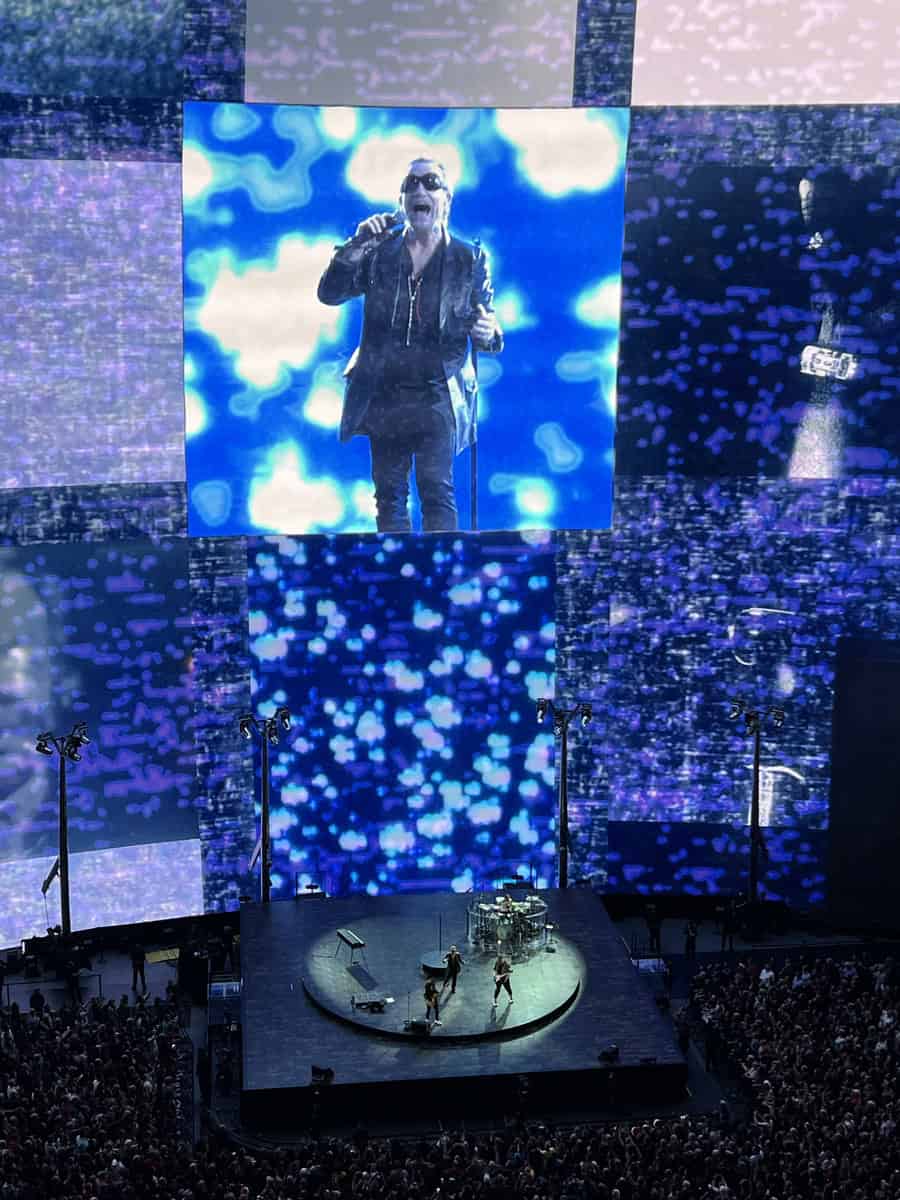 U2 performs on a circular stage at The Sphere in Las Vegas, with a massive digital screen behind them displaying a close-up of Bono singing in front of a blue and white swirling background. The crowd surrounds the stage, and the visuals fill the towering walls of the venue.