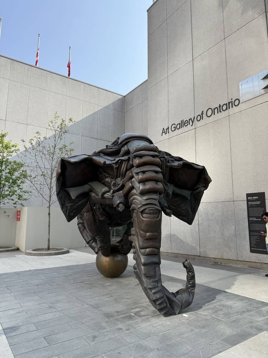 Large bronze elephant sculpture balanced on a sphere outside the Art Gallery of Ontario. The piece is abstract and powerful, positioned in a minimalist courtyard with concrete walls and subtle signage on the building.