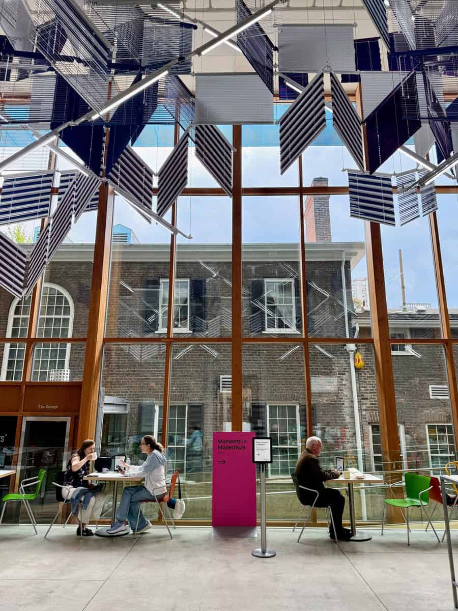 Bright seating area inside the Art Gallery of Ontario, with visitors chatting over coffee beside large glass windows that frame the historic Grange House outside. Overhead, a hanging geometric art installation made of striped slats adds a bold, modern touch.