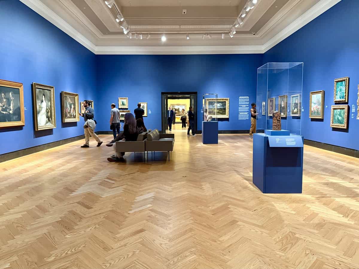 Spacious room at the AGO with vivid blue walls and parquet wood flooring, displaying European master paintings in ornate frames. Visitors stroll and sit while taking in the diverse collection of portraits and impressionist works.