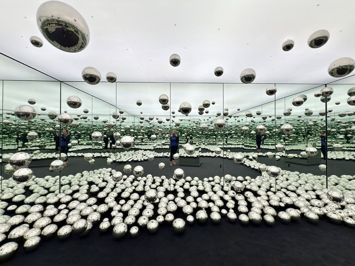 Immersive mirrored room filled with metallic spheres both suspended in air and covering the floor, reflecting endlessly in all directions. A person can be seen taking a photo, adding scale to the surreal and futuristic space.