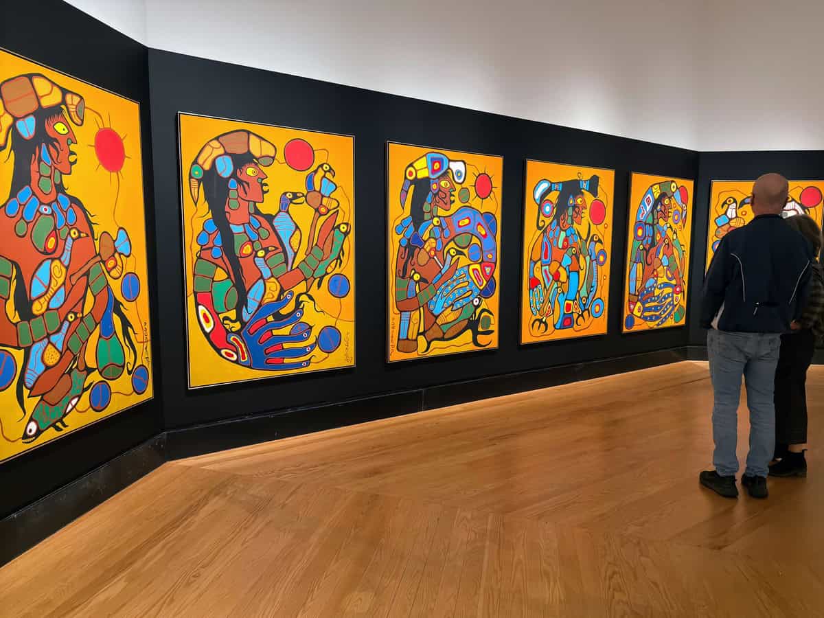 Gallery wall at the Art Gallery of Ontario featuring a vibrant series of paintings by Norval Morrisseau, known for his bold lines and vivid, symbolic depictions of Indigenous stories. Two visitors stand observing the works, which are set against a black backdrop with a warm wood floor below.