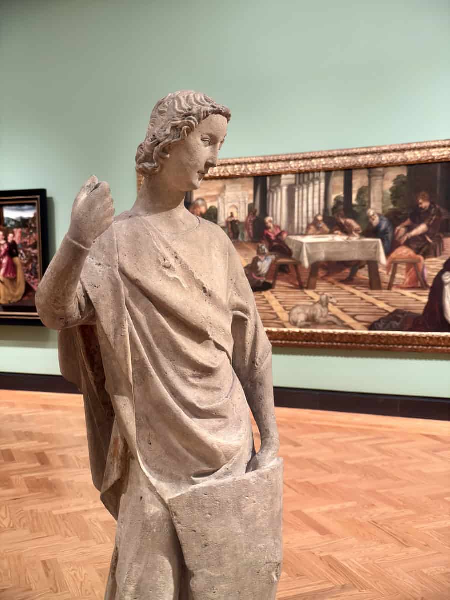 Close-up of a weathered classical stone sculpture of a female figure in front of a richly detailed Renaissance painting depicting a banquet scene. The light green wall and ornate parquet flooring frame the artworks in a traditional yet serene gallery space.