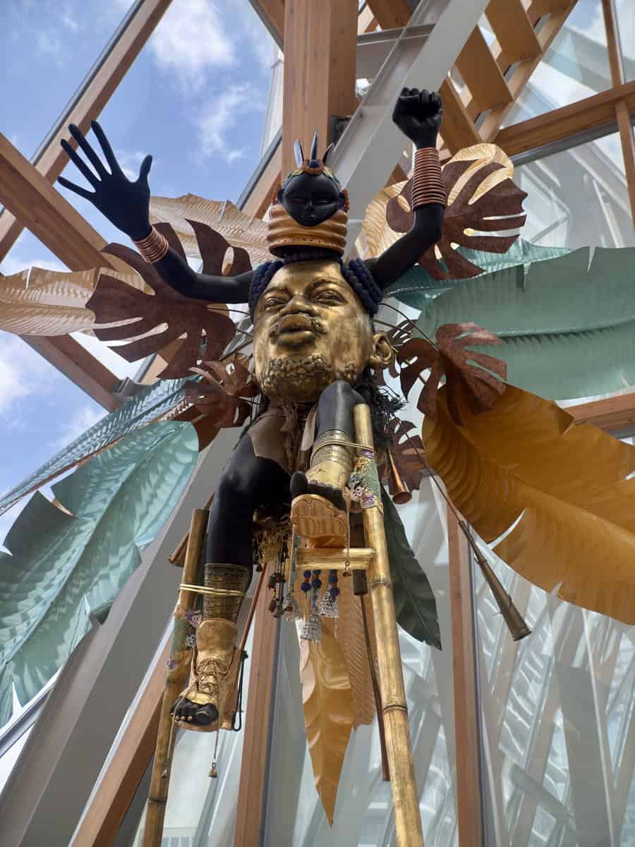 Dramatic sculpture inside the Art Gallery of Ontario featuring a gold-painted face with a black figure on top, surrounded by large stylized tropical leaves. The installation is suspended in an atrium with wooden beams and glass walls, giving it a striking presence against the sky.