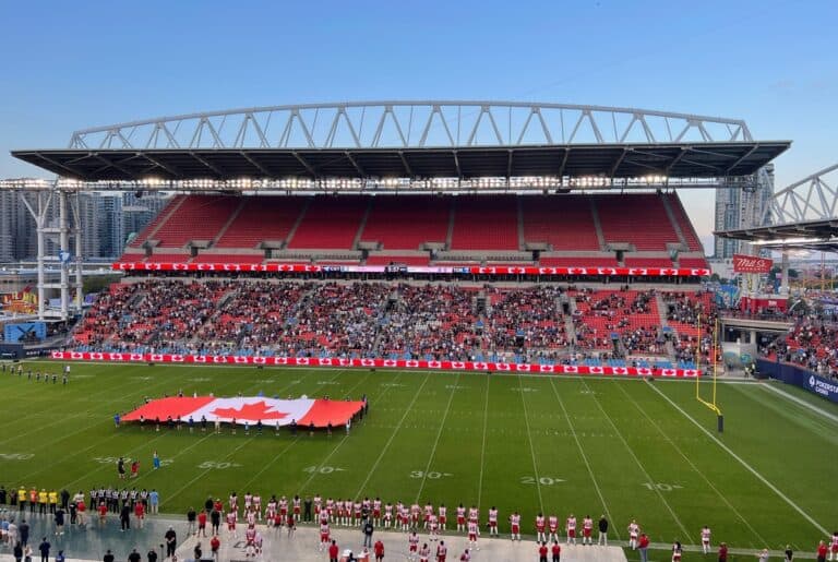 Panoramic view of BMO Field showing a large Canadian flag being held across the field before kickoff, as teams line up along the sidelines. Digital displays around the stadium are lit with Canadian flags, and fans in the stands watch the pre-game ceremony under a clear evening sky.