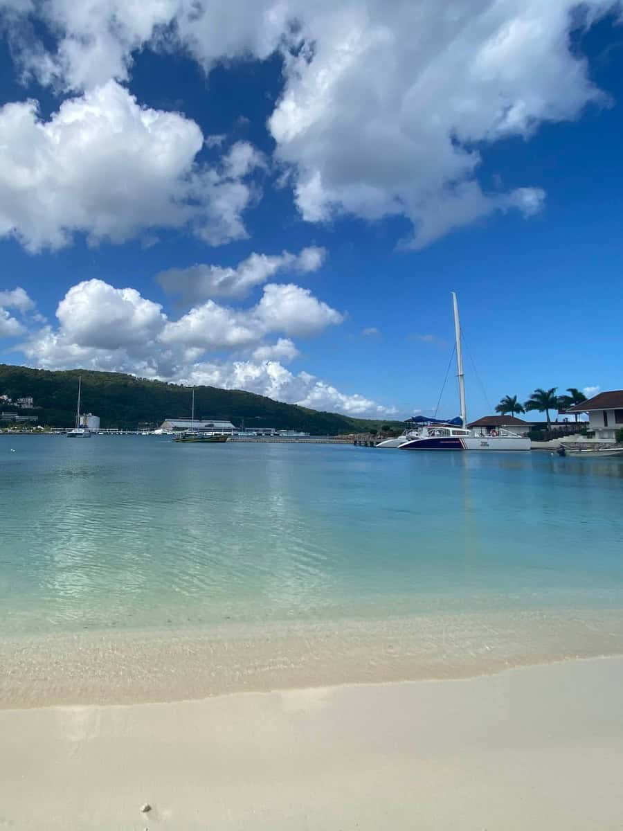 Tranquil beach scene with powdery white sand and crystal-clear water at Moon Palace Jamaica. A catamaran floats near the shore under a partly cloudy sky, with green hills in the distance.