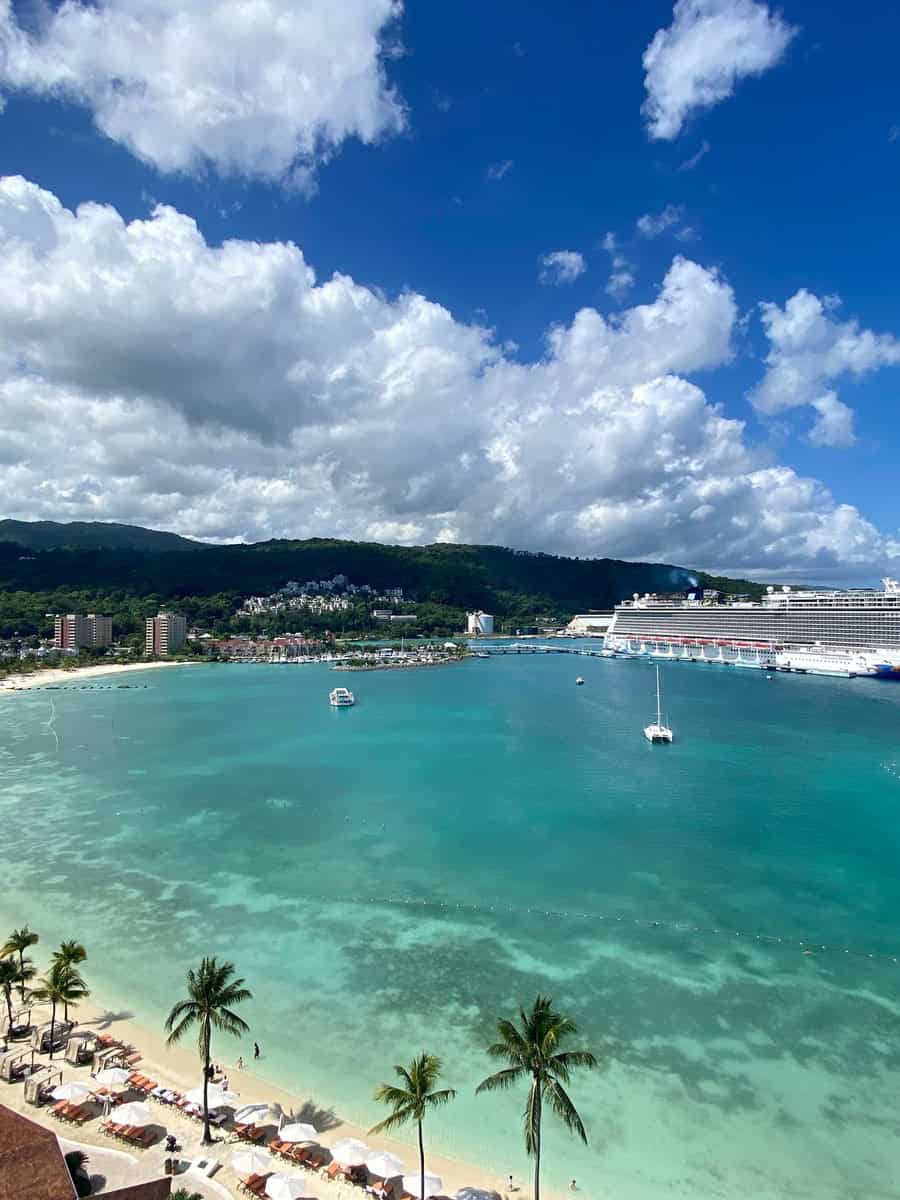 A stunning view of the turquoise Caribbean Sea with palm trees and beach umbrellas lining the golden sands of Moon Palace Jamaica. A large cruise ship is docked nearby under a vibrant blue sky with fluffy clouds.