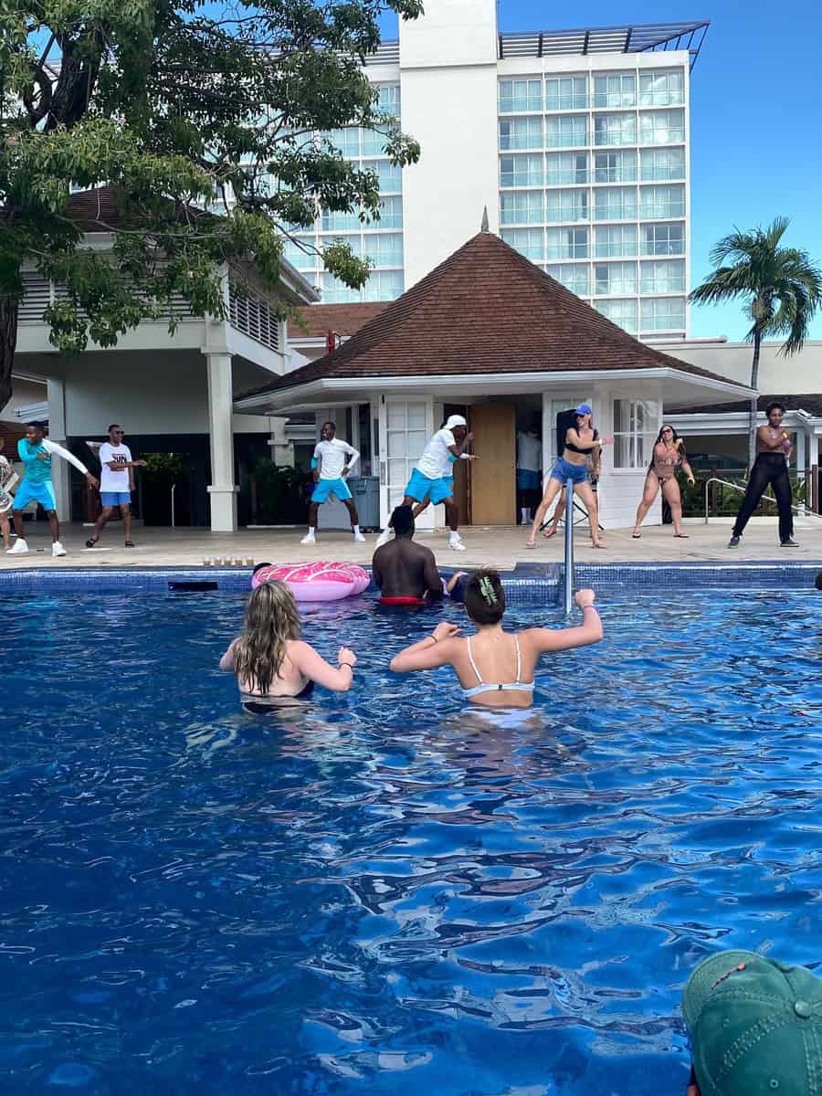 Energetic poolside dance activity led by Moon Palace Jamaica staff in turquoise outfits, with resort guests participating from the pool. The scene is framed by tropical trees and resort buildings.
