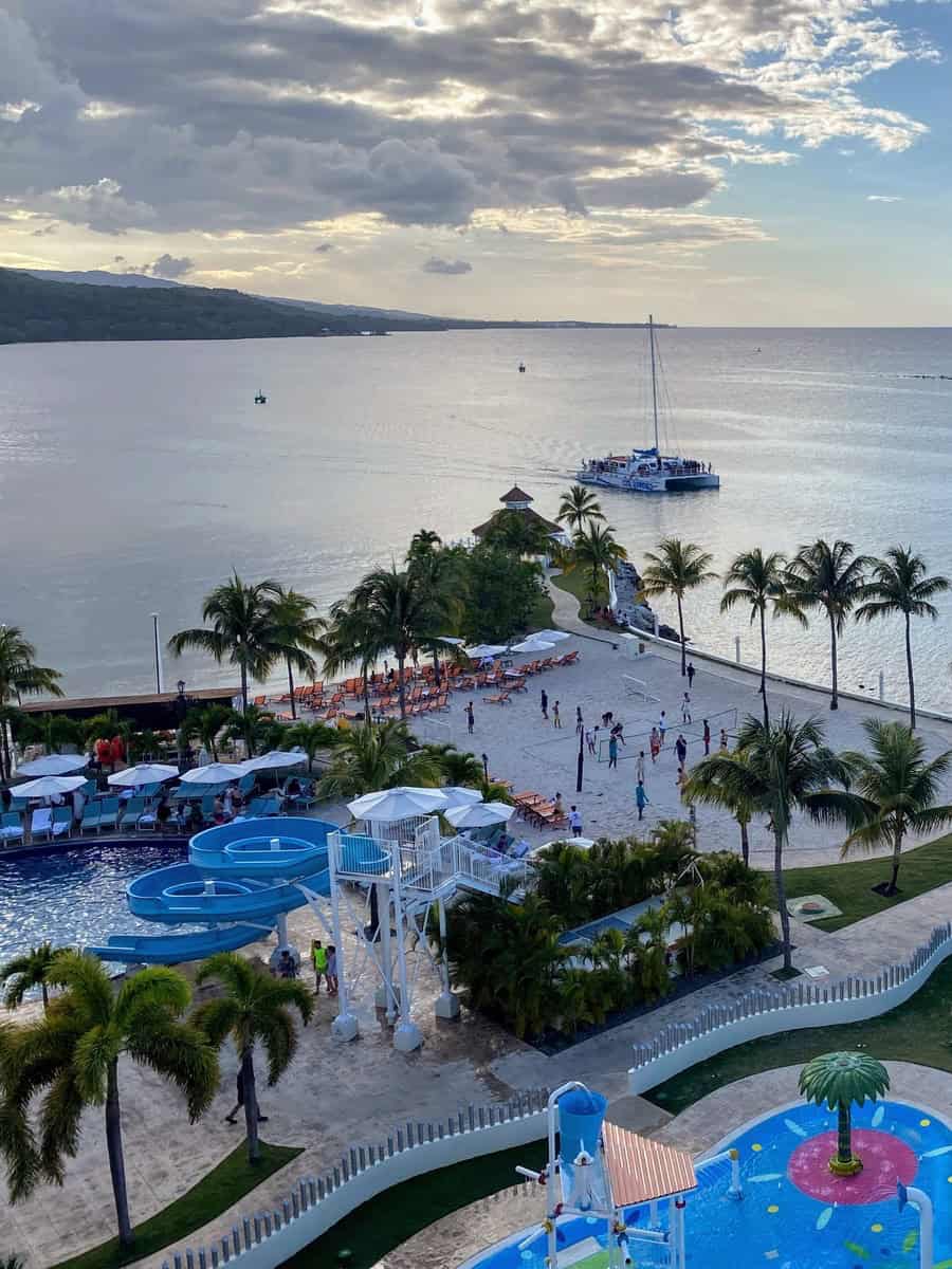 Overhead view of the resort’s pools, splash pad, and beach volleyball area at Moon Palace Jamaica. A catamaran sails in the background as guests enjoy water slides and lounge chairs along the beach.