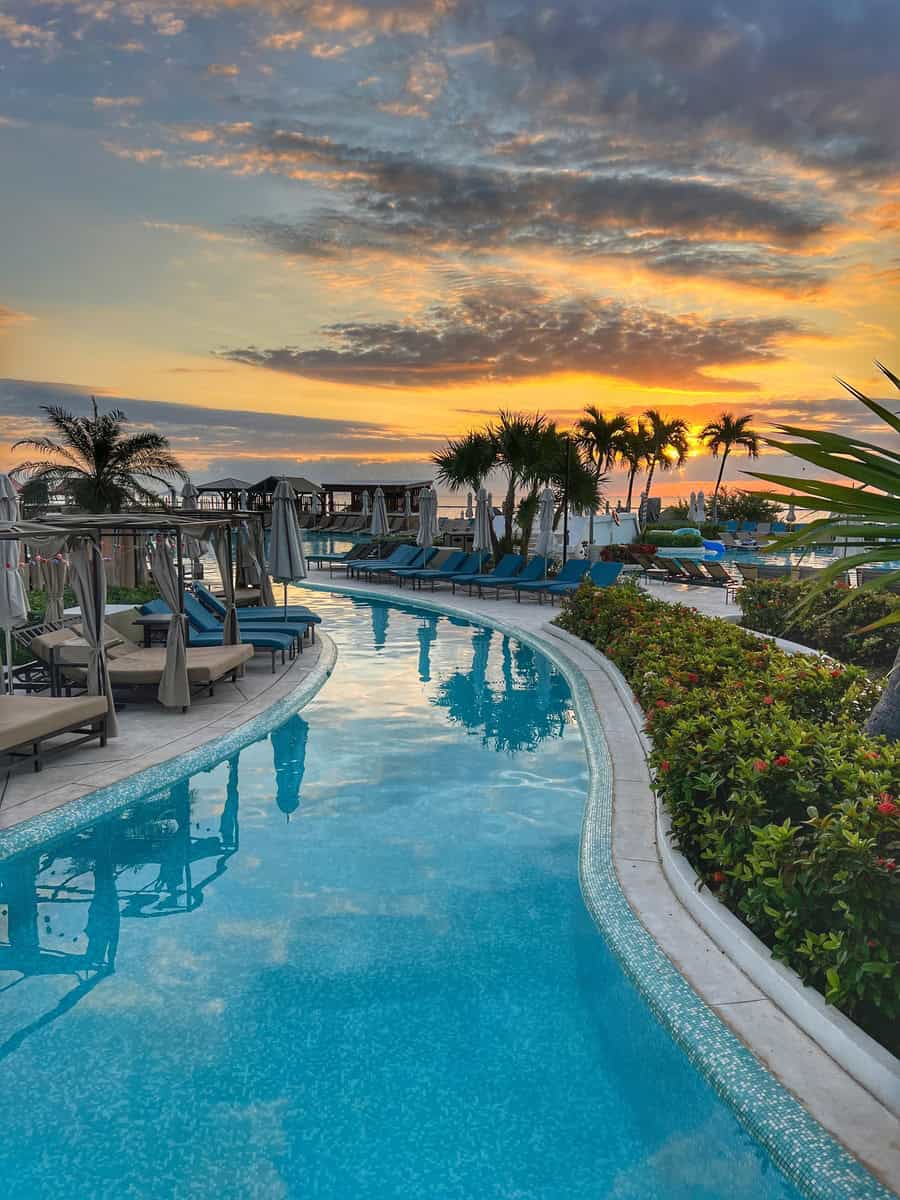 A serene view of a winding pool at Moon Palace Cancun just after sunrise, with soft reflections of palm trees and cabanas in the water. The sky glows in hues of orange and pink, casting a tranquil light over the peaceful pool area.