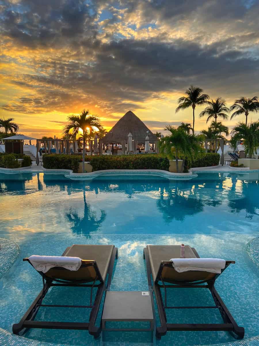 Two poolside lounge chairs face a vibrant sunrise at Moon Palace Cancun, with a thatched-roof palapa, palm trees, and dramatic clouds reflecting in the pool. The tranquil morning scene invites relaxation and quiet moments.