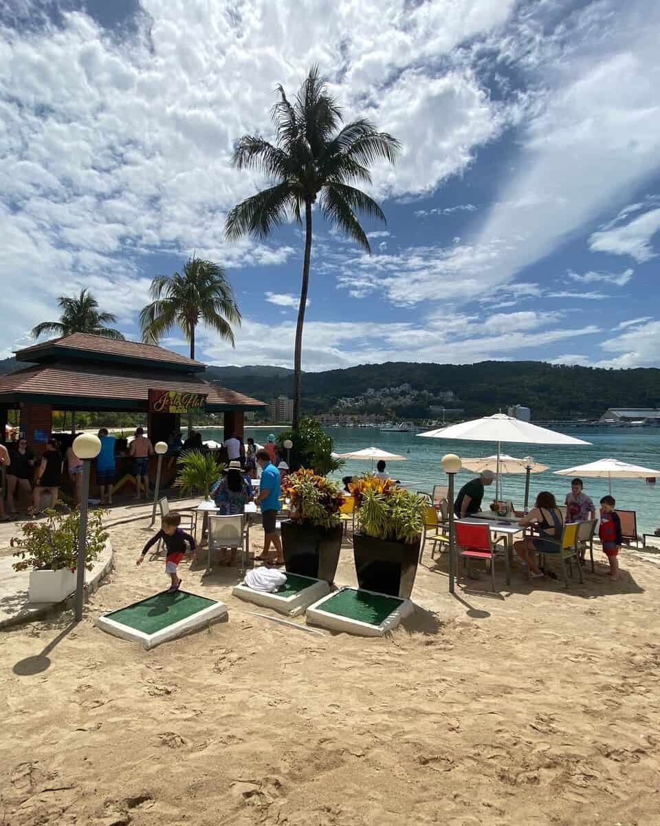Guests gather at the lively Jerk Hut on the beach at Moon Palace Jamaica, surrounded by tropical plants and colorful beach chairs. Palm trees sway under a partly cloudy sky, while the turquoise water and hills of Ocho Rios provide a scenic Caribbean backdrop.