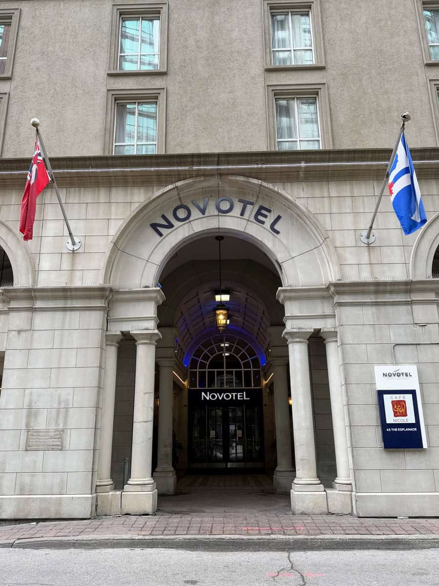 Front view of the Novotel Toronto Centre hotel, featuring a stone archway entrance flanked by two flags—Ontario’s provincial flag and the City of Toronto flag. The arched glass doors lead into a softly lit lobby, with signage for Café Nicole displayed on the right.