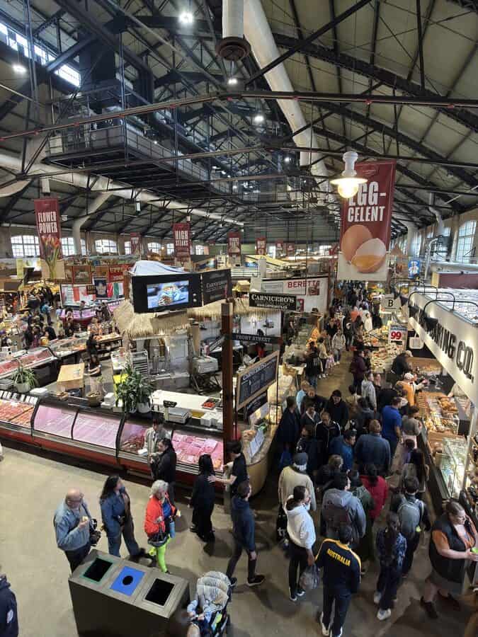Bustling scene inside St. Lawrence Market in Toronto, with crowds exploring a variety of food vendors under a high industrial ceiling with hanging red banners.