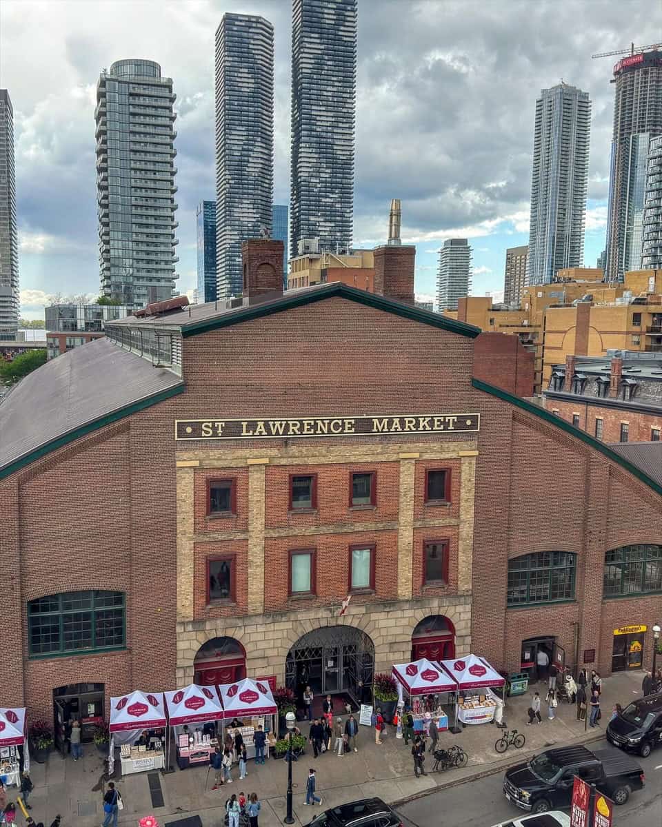 View of the historic red-brick St. Lawrence Market building in Toronto, with vendor tents set up along the sidewalk and people browsing stalls. Tall modern condos tower in the background under a dramatic cloudy sky, creating a contrast between old and new architecture.
