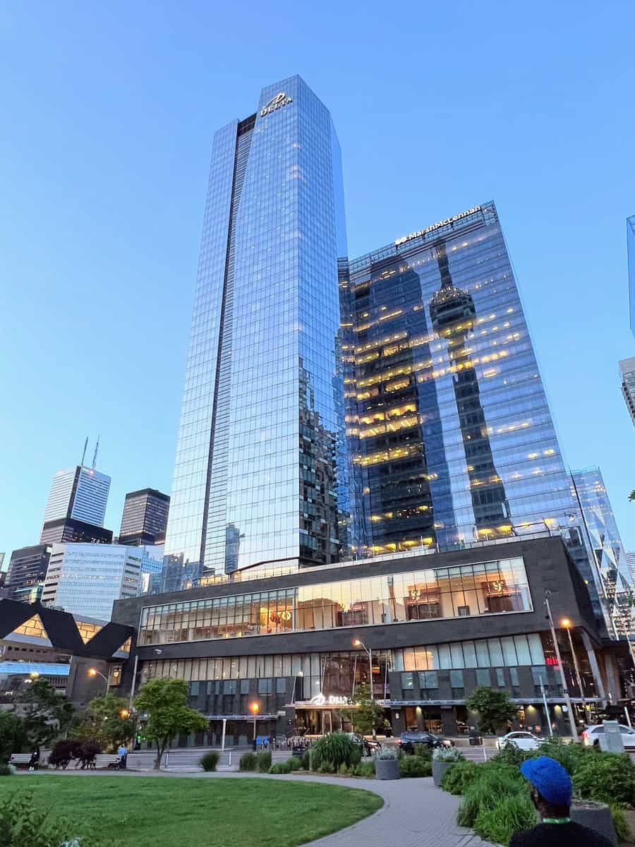 Tall glass skyscraper of the Delta Hotel in downtown Toronto, reflecting the CN Tower and nearby high-rises at dusk. A grassy park with a walking path and a person in a blue cap are visible in the foreground, adding depth and contrast to the sleek urban scene.