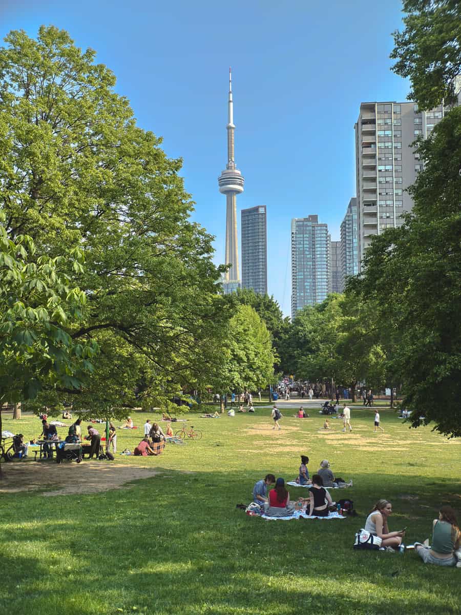Lively summer scene at Grange Park in Toronto with groups of people lounging on the grass, picnicking, and cycling. Towering trees frame a clear view of the CN Tower and surrounding high-rises in the distance under a bright blue sky.