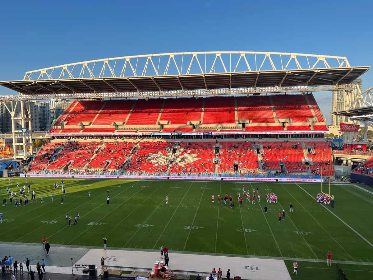 Wide view of BMO Field in Toronto during a football game warm-up, with players scattered across the field and fans beginning to fill the bright red stadium seats. The late afternoon sun casts long shadows across the turf, highlighting the field markings and "CFL" logos near the sidelines.