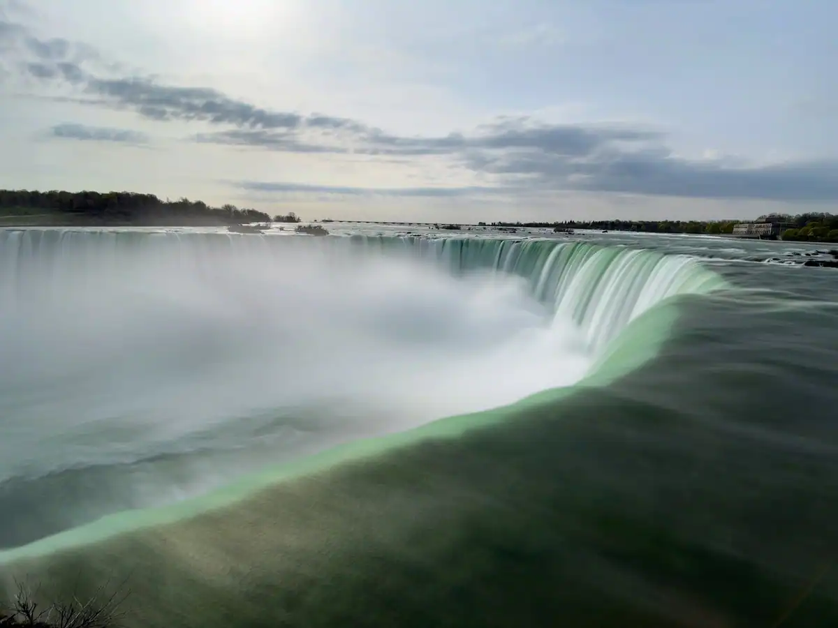 Stunning early morning shot of Niagara’s Canadian Horseshoe Falls, with emerald green water curving dramatically before plunging down, framed by a serene sky and distant treeline