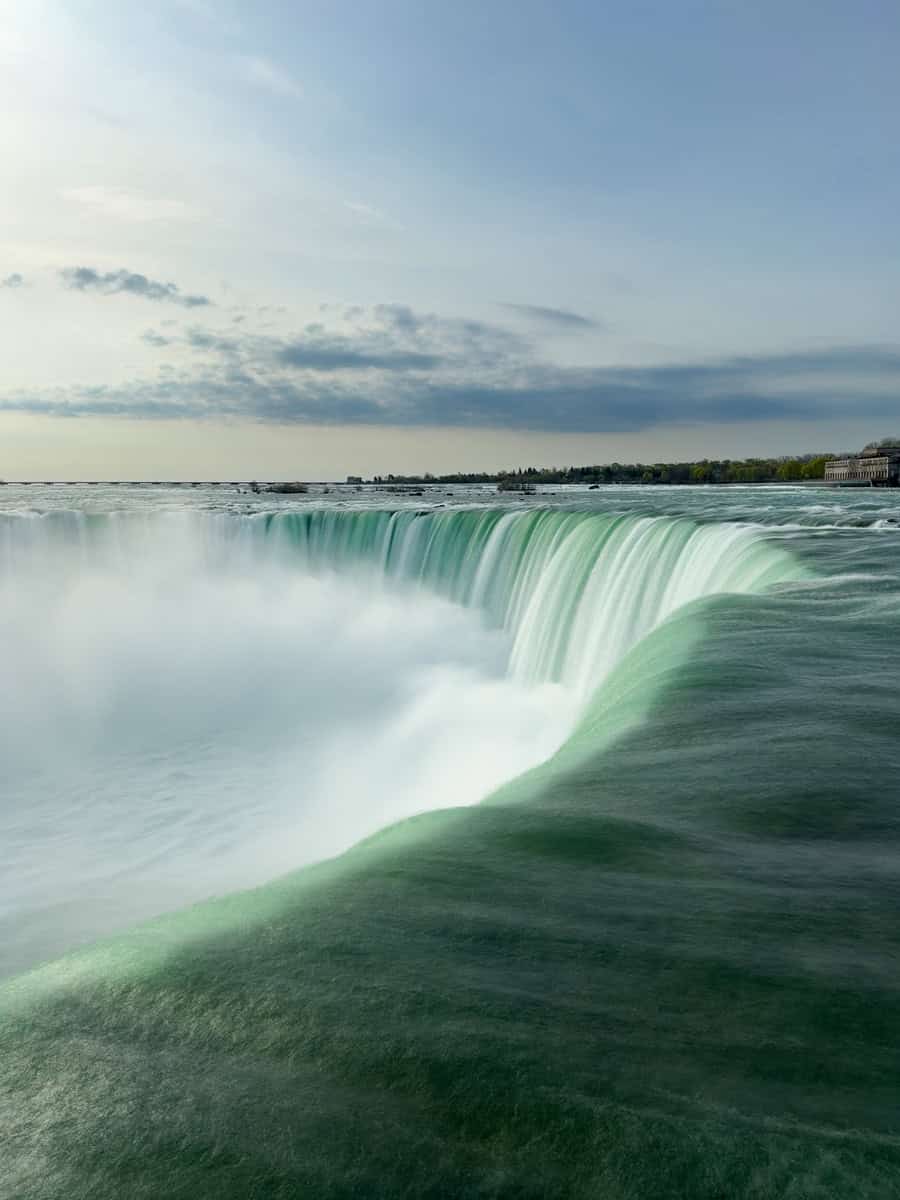 Soft, misty view from the brink of Niagara Falls, Ontario, Canada in the early morning, showcasing smooth, flowing water cascading powerfully over the edge under a cloudy sky.