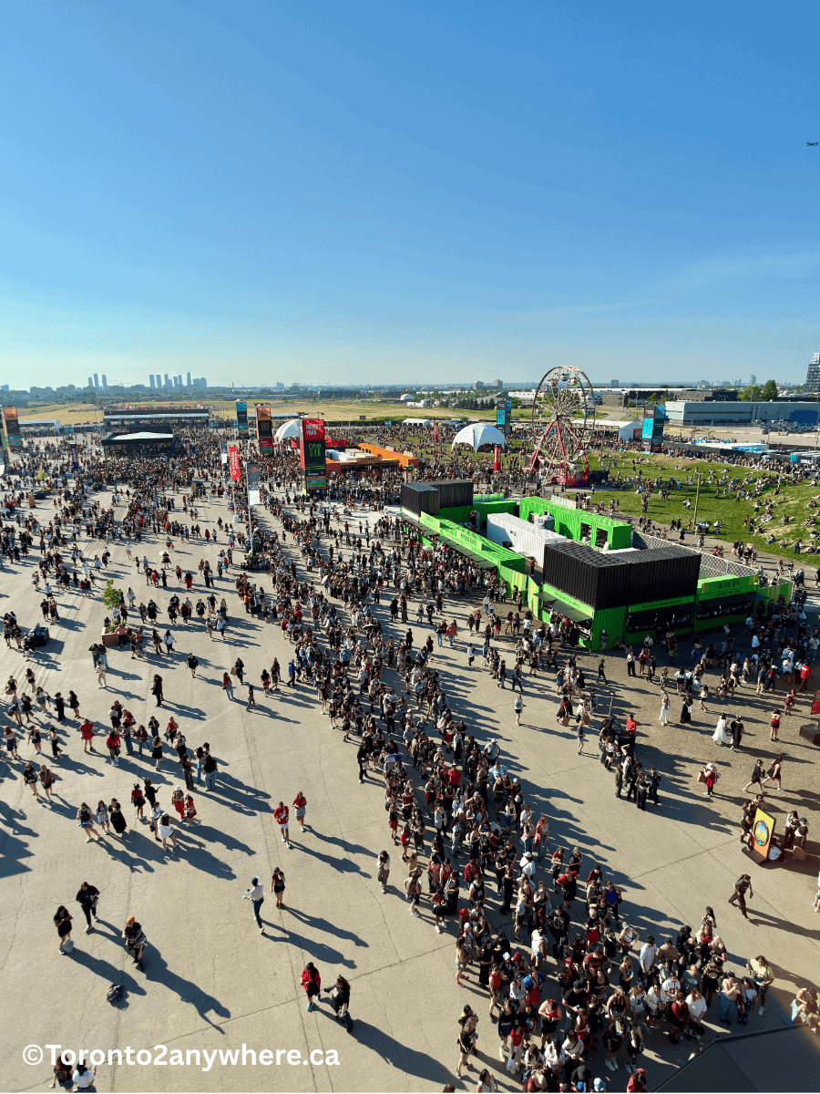 A wide aerial view of fans arriving to a summer concert at the Rogers Stadium at Downsview airport in Toronto. Large lines are visible for merchandise, food and a Ferris wheel on a sunny day. People are lined up along wide concrete paths, and the city skyline is faintly visible in the distance.